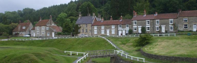 Stone cottages above a village green with a stream and a small footbridge, with wooded hills behind
